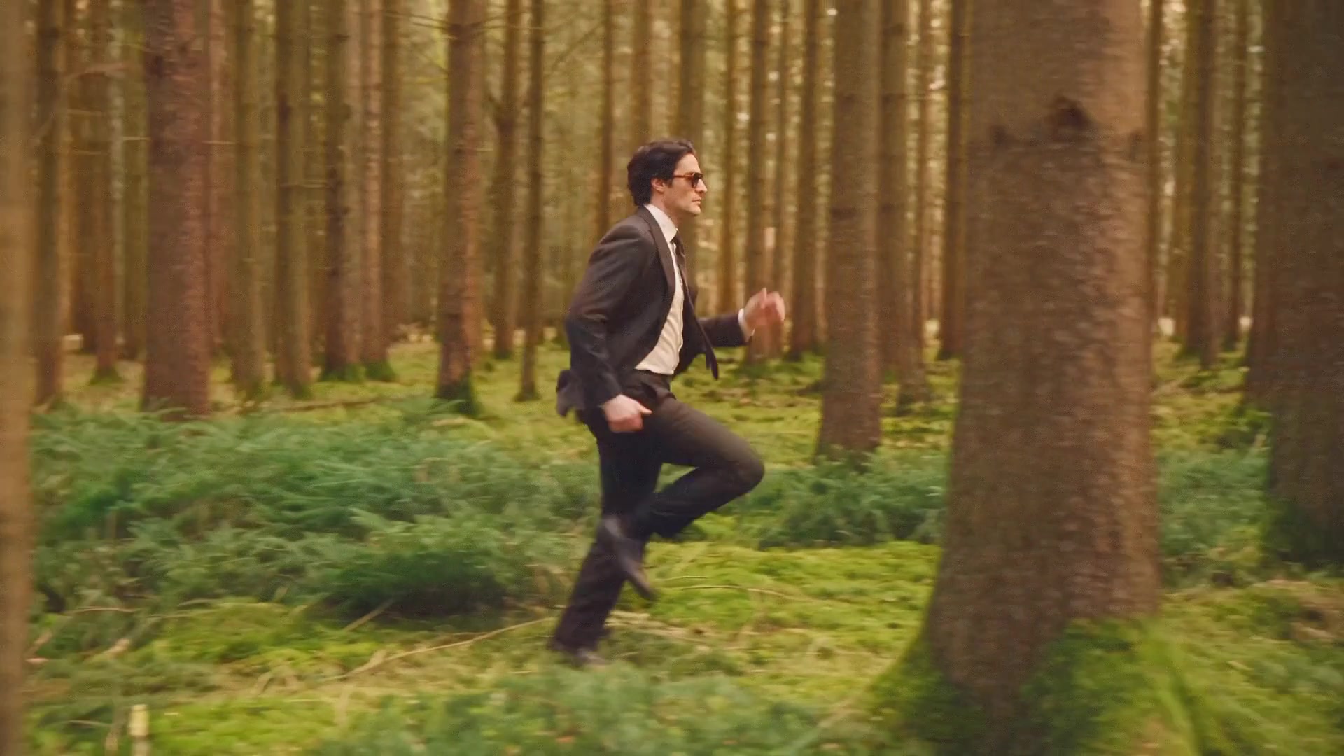 A man in a dark brown suit runs through a pine forest, the camera tracking beside him as trunks sweep past.
