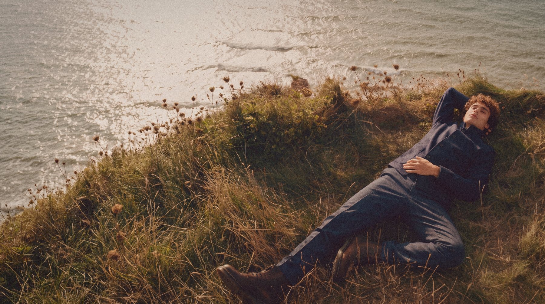 A man in a navy shirt jacket lies in the grass at a cliff edge above the sea.