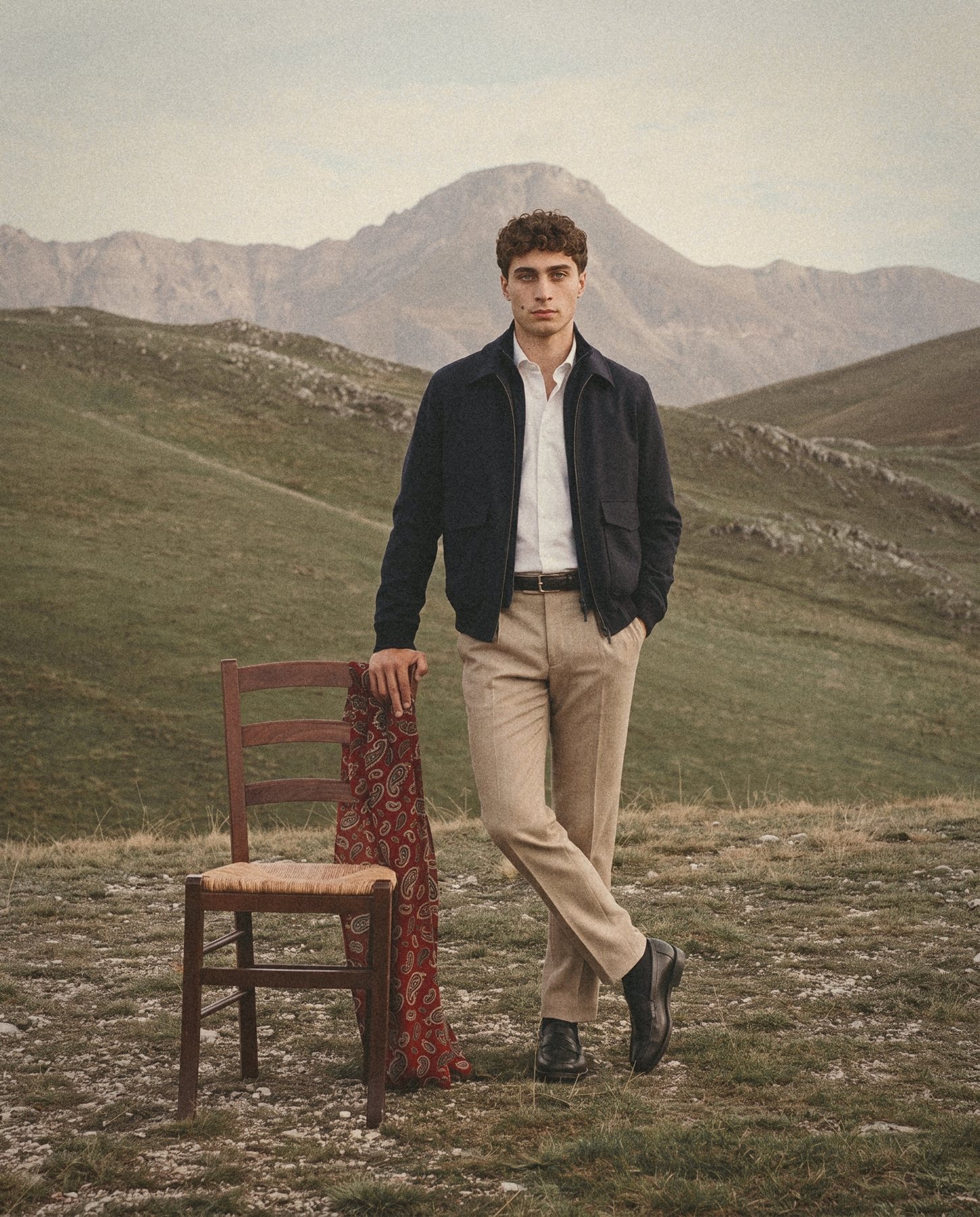 A man in a navy zip jacket and sand trousers stands beside a rush seat chair on a mountain plateau.