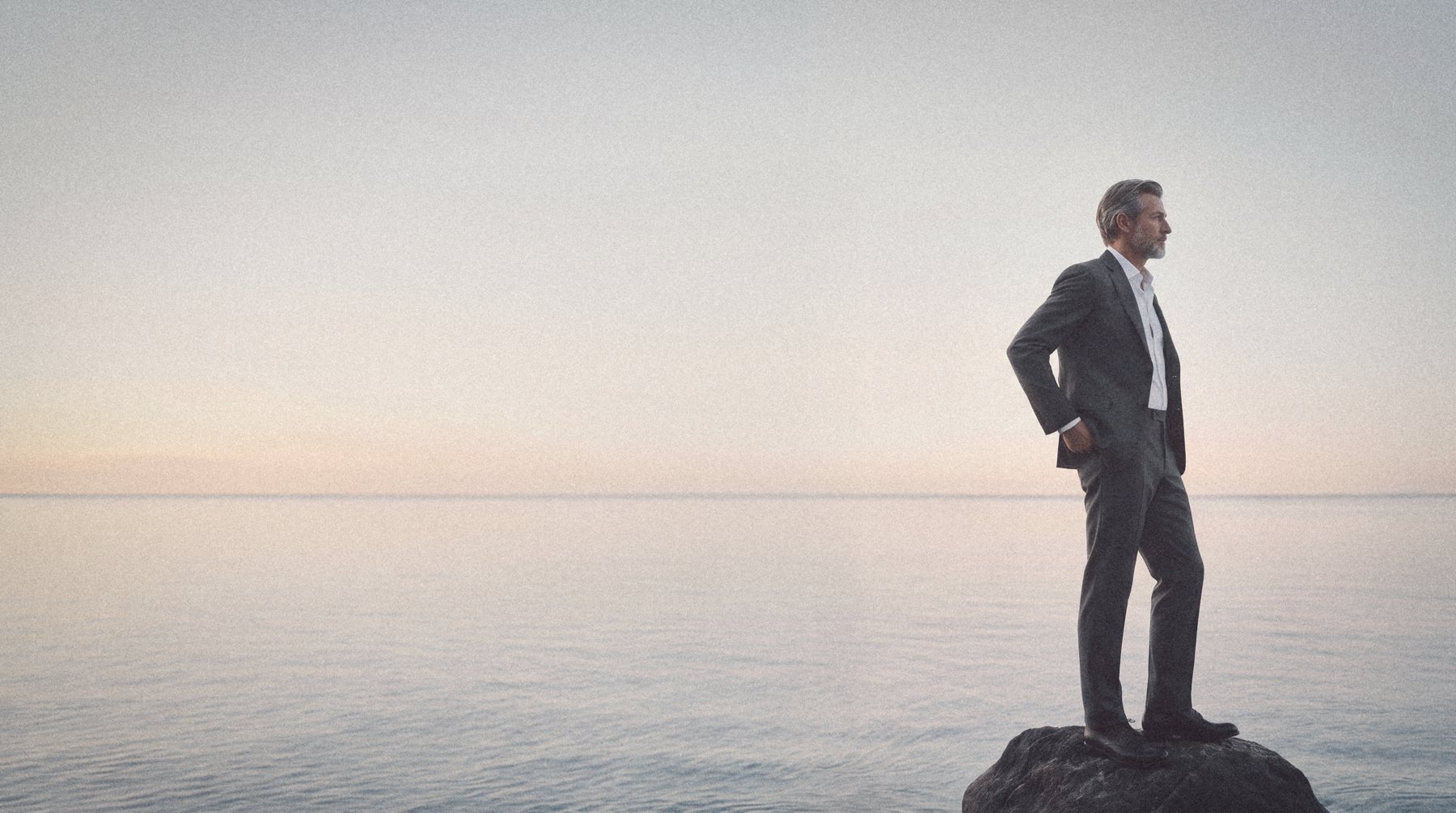 A grey haired man in a charcoal suit stands on a rock above a still sea at blue hour, looking out.