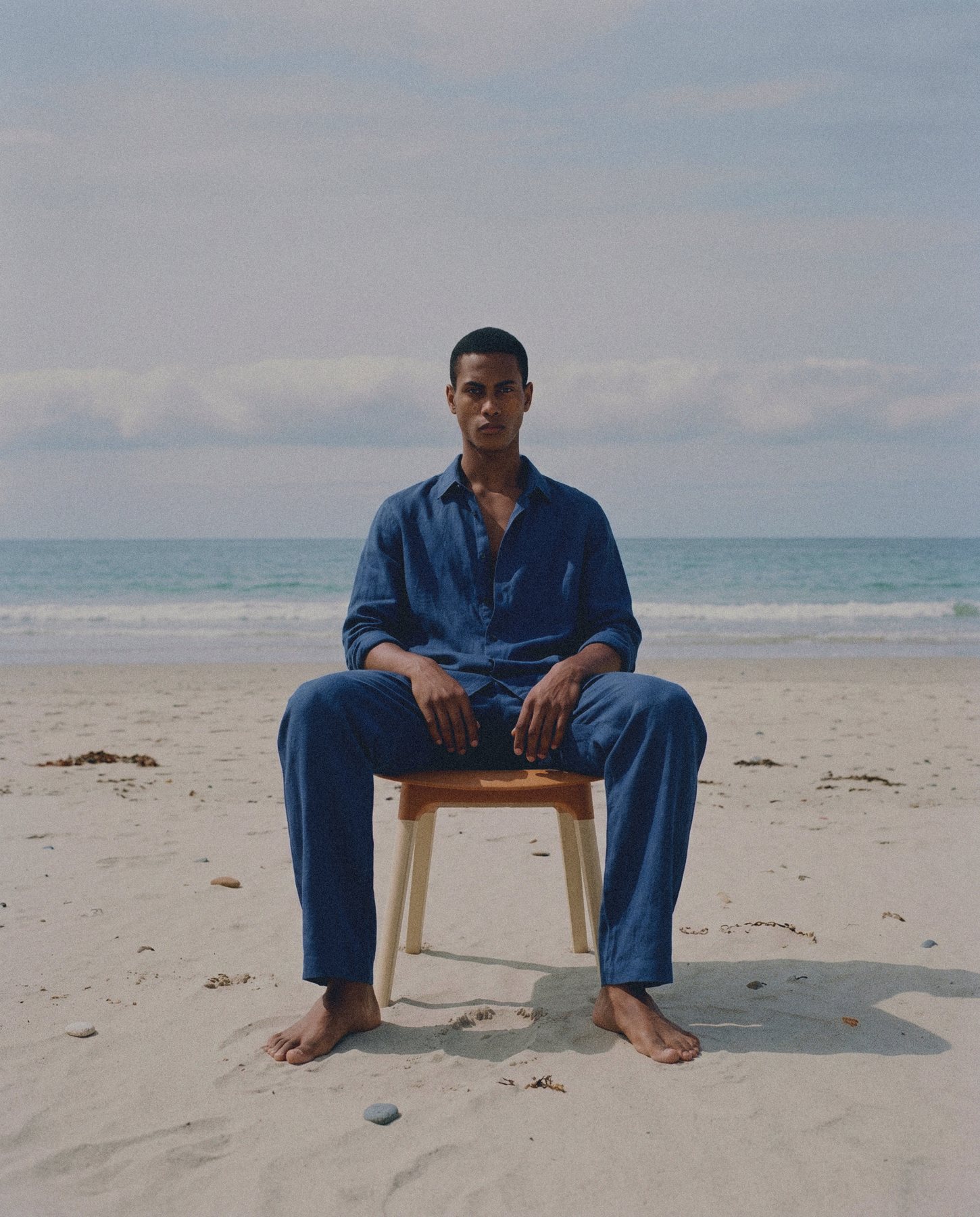 A man in a blue linen shirt and trousers sits barefoot on a wooden stool on a beach.