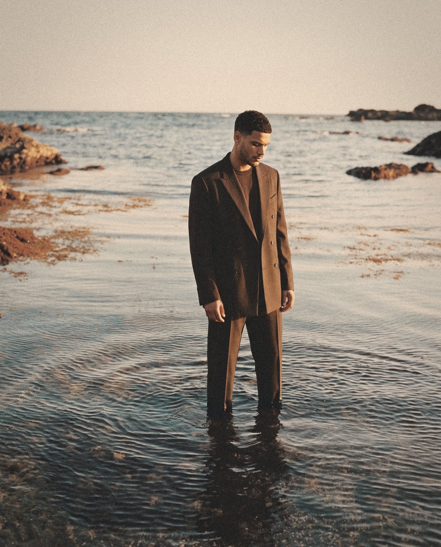 A man in a brown double breasted suit stands in shallow water at golden hour, looking down.