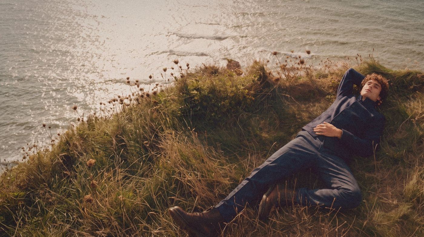 A man in a navy shirt jacket lies in the grass at a cliff edge above the sea.