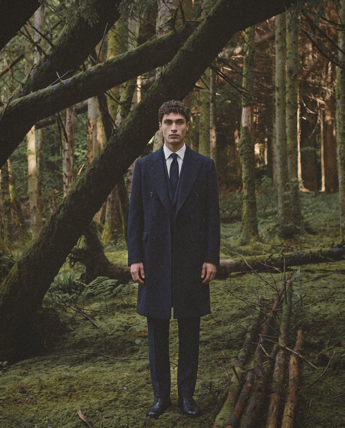 A man in a navy double breasted overcoat stands in a mossy forest.